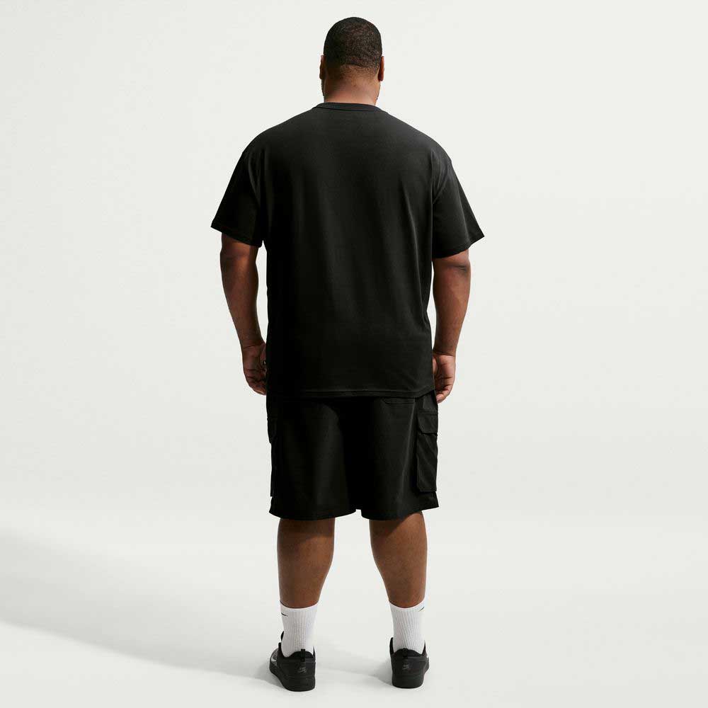 A man stands facing away from the camera against a plain white background, wearing a black Nike SB Cell Phone Tee (IM4424010), paired with black shorts, white socks, and black shoes. The T-shirt features sweat-wicking fabric to keep him comfortable while active.