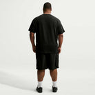 A man stands facing away from the camera against a plain white background, wearing a black Nike SB Cell Phone Tee (IM4424010), paired with black shorts, white socks, and black shoes. The T-shirt features sweat-wicking fabric to keep him comfortable while active.