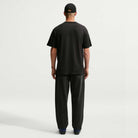 A person stands facing away, wearing a black Nike SB Cell Phone Tee (IM4424010) paired with dark loose pants, a black cap, and dark shoes, on a plain white background. The sweat-wicking Nike SB Skateboarding t-shirt combines comfort and style for an effortlessly cool look.
