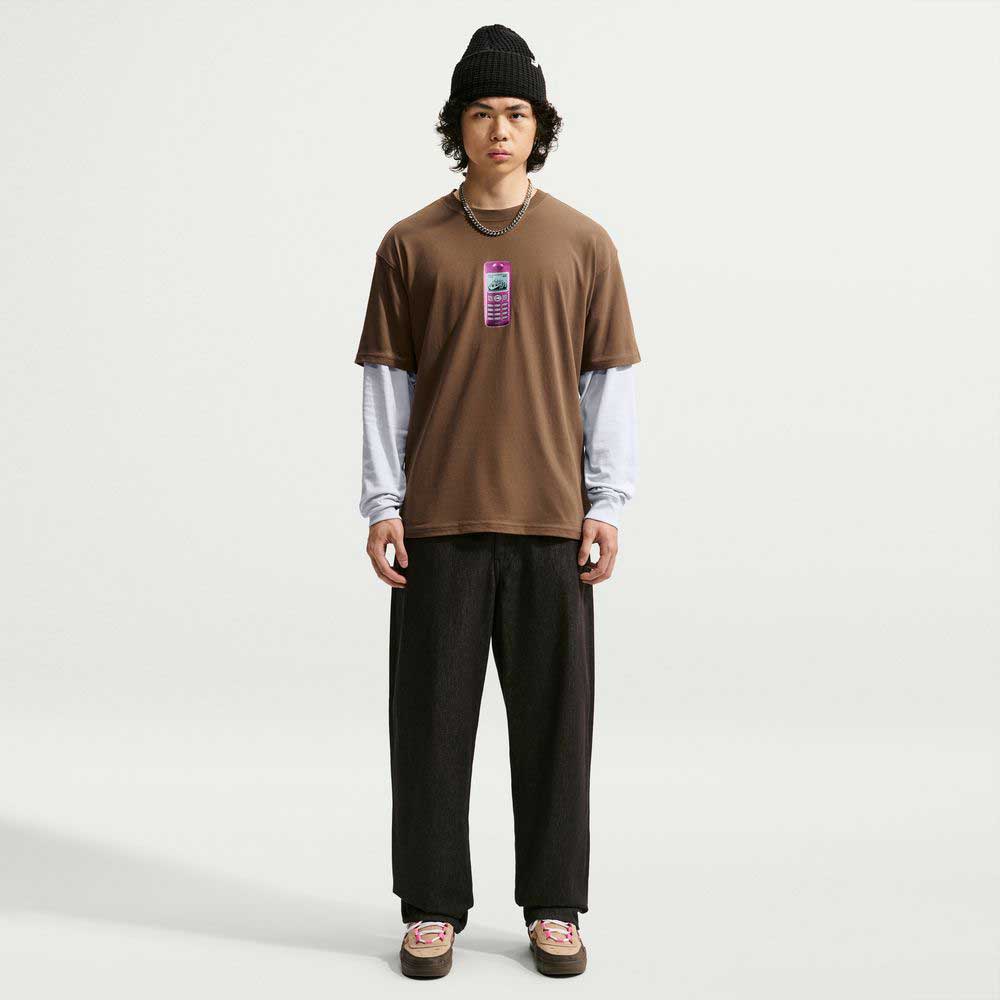 A person wearing a black beanie, a Cell Phone Tee Brown IM4424235 T-Shirt from Nike SB Skateboarding over a white long sleeve, black pants, and brown shoes stands facing forward against a plain white background. The sweat-wicking skateboarding tee adds both style and comfort to the look.