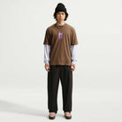 A person wearing a black beanie, a Cell Phone Tee Brown IM4424235 T-Shirt from Nike SB Skateboarding over a white long sleeve, black pants, and brown shoes stands facing forward against a plain white background. The sweat-wicking skateboarding tee adds both style and comfort to the look.