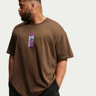 A man in the brown Cell Phone Tee (IM4424235) from Nike SB Skateboarding, featuring a graphic of a purple cell phone on the chest, stands against a plain, light background and looks to his left. This comfortable, sweat-wicking T-shirt effortlessly blends style and performance for everyday wear.
