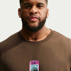 Wearing the brown Nike SB Cell Phone Tee (IM4424235), a man stands against a plain background, styled with earrings and holding a purple device at chest level that features a Nike SB sticker. The skateboarding t-shirt’s sweat-wicking design enhances his laid-back look.
