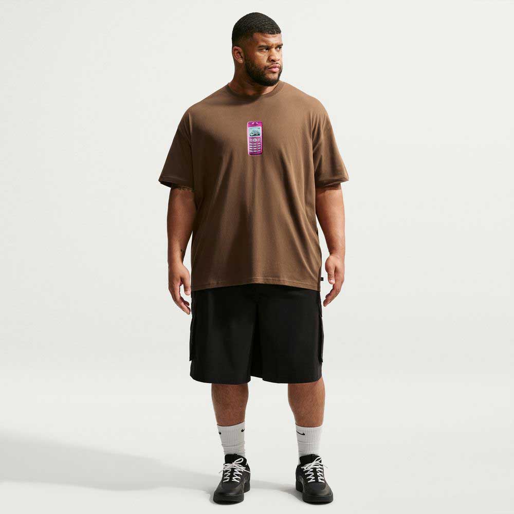 A man stands against a plain white background, wearing a loose brown Cell Phone Tee Brown IM4424235 T-Shirt from Nike SB Skateboarding, which features a vertical graphic on the chest. He pairs the tee with black shorts, white socks, and black sneakers. The sweat-wicking skateboarding top keeps him comfortable as he looks slightly to his left with his arms relaxed.