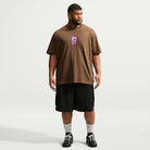 A man stands against a plain white background, wearing a loose brown Cell Phone Tee Brown IM4424235 T-Shirt from Nike SB Skateboarding, which features a vertical graphic on the chest. He pairs the tee with black shorts, white socks, and black sneakers. The sweat-wicking skateboarding top keeps him comfortable as he looks slightly to his left with his arms relaxed.