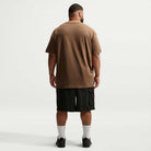 A man stands facing away from the camera, wearing a brown Nike SB Cell Phone Tee (Style: IM4424235)—a premium skateboarding T-shirt—paired with black shorts, white socks, and black shoes against a plain white background.