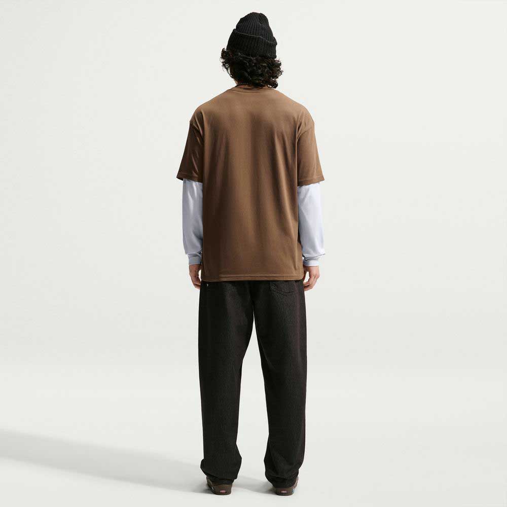 A person stands facing away, wearing a black beanie and loose dark pants, with the Nike SB Cell Phone Tee Brown IM4424235 Skateboarding T-Shirt layered over a white long-sleeve shirt. The sweat-wicking skate tee stands out against the plain, light background.