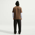 A person stands facing away, wearing a black beanie and loose dark pants, with the Nike SB Cell Phone Tee Brown IM4424235 Skateboarding T-Shirt layered over a white long-sleeve shirt. The sweat-wicking skate tee stands out against the plain, light background.