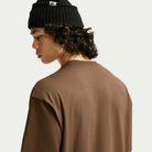 A person with curly hair, seen in side profile and turned away from the camera against a plain background, is wearing a black knit beanie and the Nike SB Skateboarding Cell Phone Tee Brown IM4424235 t-shirt.
