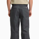 A person wearing Charcoal Loose Fit Work Pant A88KB0CH by Dickies Skateboarding, made from durable twill, viewed from the back. Features two flap-back pockets, a logo patch above the right pocket, and a neatly tucked-in beige shirt.