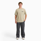 A young person faces forward, wearing a beige tee, white sneakers, and Charcoal Loose Fit Work Pant A88KB0CH Dickies Skateboarding made from durable twill fabric. The background is plain white.