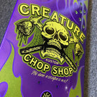 Close-up of the Chop Shop Flame 8.50in x 32.05in Creature Skateboard Shaped Deck, made from 7-ply North American maple with a yellow skull, crossed axes, spider webs, green flames, and CREATURE CHOP SHOP KUSTOM graphic. Text: No one escapes us!.