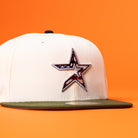 The Stars Over Houston UH New Era Hat 5950 Fitted features a cream color, green brim, and a star logo with a U.S. flag pattern—perfect for adding bold style to any Houston Cougars fan’s collection. Displayed on a bright orange background.