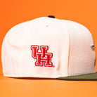 The Stars Over Houston UH New Era Hat 5950 Fitted, featuring a white crown, green brim, and the red and white Houston Cougars logo embroidered on the side, is displayed against an orange background.