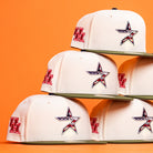 A stack of Stars Over Houston UH New Era Hat 5950 Fitted hats in white with olive brims, a red UH logo on the side, and a front star outlined in black with an American flag pattern—all on an orange background.