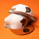 Two white mesh The Wrangler Chrome Rust Sugarland Space Cowboys New Era Hat 5950 Fitted caps with orange brims and a cartoon cowboy logo are stacked on an orange surface. One cap features a Championship Pop Fly patch on the side, capturing the playful spirit of a Space Cowboys hat. These designs embody Chrome Rust Champ 24 vibes, giving classic style a fresh, modern edge.
