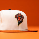 Introducing The Wrangler Chrome Rust Sugarland Space Cowboys New Era Hat 5950 Fitted—a white and orange trucker cap featuring an embroidered patch of a strawberry sporting a cowboy hat and bandana against an orange background. This lively design embodies the energetic spirit of the Space Cowboys Hat collection, making it ideal for anyone eager to add some playful flair to their collection.