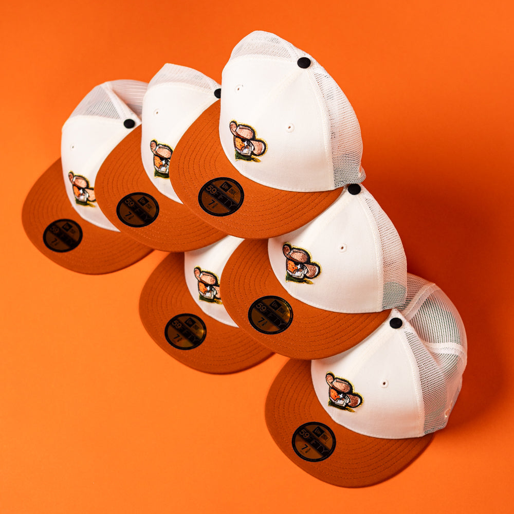 Six “The Wrangler Chrome Rust Sugarland Space Cowboys New Era Hat 5950 Fitted” trucker hats—featuring a white and orange color scheme with a cartoon bear patch—are arranged in a triangular formation on an orange background. Each hat showcases mesh backs and black sticker labels on the brims, combining classic style with modern flair reminiscent of the lively Space Cowboys aesthetic.