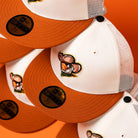 Stacked white and orange The Wrangler Chrome Rust Sugarland Space Cowboys New Era Hat 5950 Fitted trucker hats, each with mesh backs and a cartoon cowboy potato embroidered on the front panel, are displayed against an orange background—a vibrant showcase of Space Cowboys style and playful flair.