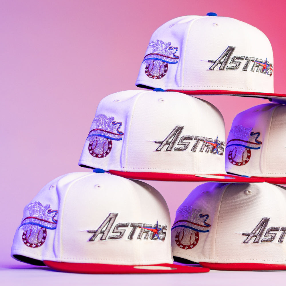 A pyramid of The Diplomat Chrome Scarlet Houston Astros New Era Hat 5950 Fitted caps, each with red brims and white crowns, displays the sparkling Astros logo against a pink and purple gradient background.
