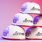 A pyramid of The Diplomat Chrome Scarlet Houston Astros New Era Hat 5950 Fitted caps, each with red brims and white crowns, displays the sparkling Astros logo against a pink and purple gradient background.