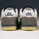 A close-up of the back of two 480 CVL New Balance Numeric x Civil Suede Leather Skateboarding Shoes shows the left shoe with “New Balance” text and the right with “CVL” text. These durable skate shoes are crafted in shades of gray, white, and cream with yellowed soles, and feature an ABZORB midsole for enhanced comfort.