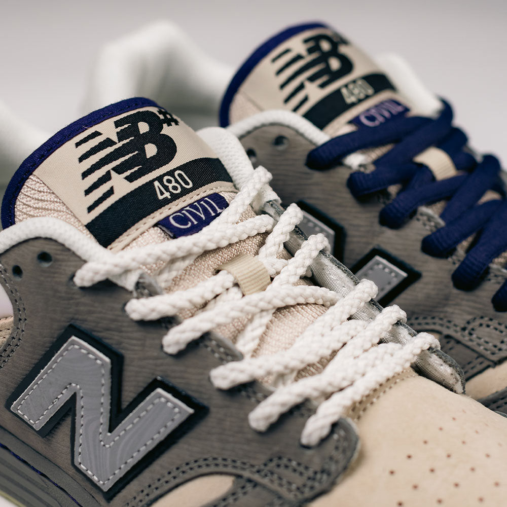 Close-up of two 480 CVL New Balance Numeric x Civil Suede Leather Skateboarding Shoes, featuring gray and beige suede uppers, white textured laces, navy blue laces on one shoe, and prominent N and NB branding on the side and tongue. Inspired by classic skate shoe silhouettes, these sneakers boast a durable design with an ABZORB midsole for enhanced comfort.