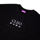The Cleo Flame Black Tee is a black cut-n-sew T-shirt featuring the CLEO logo and four white geometric star symbols beneath it. It includes a distinctive purple neck tag and is photographed laid flat on a white background, making it an essential addition to any Cleo tees collection.