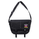 The Commute Side Bag Black by Butter Goods is made from heavy cotton ripstop and features an adjustable shoulder strap, front flap with two buckles, and a pink and yellow "BUTTER" globe logo on the lower right corner.