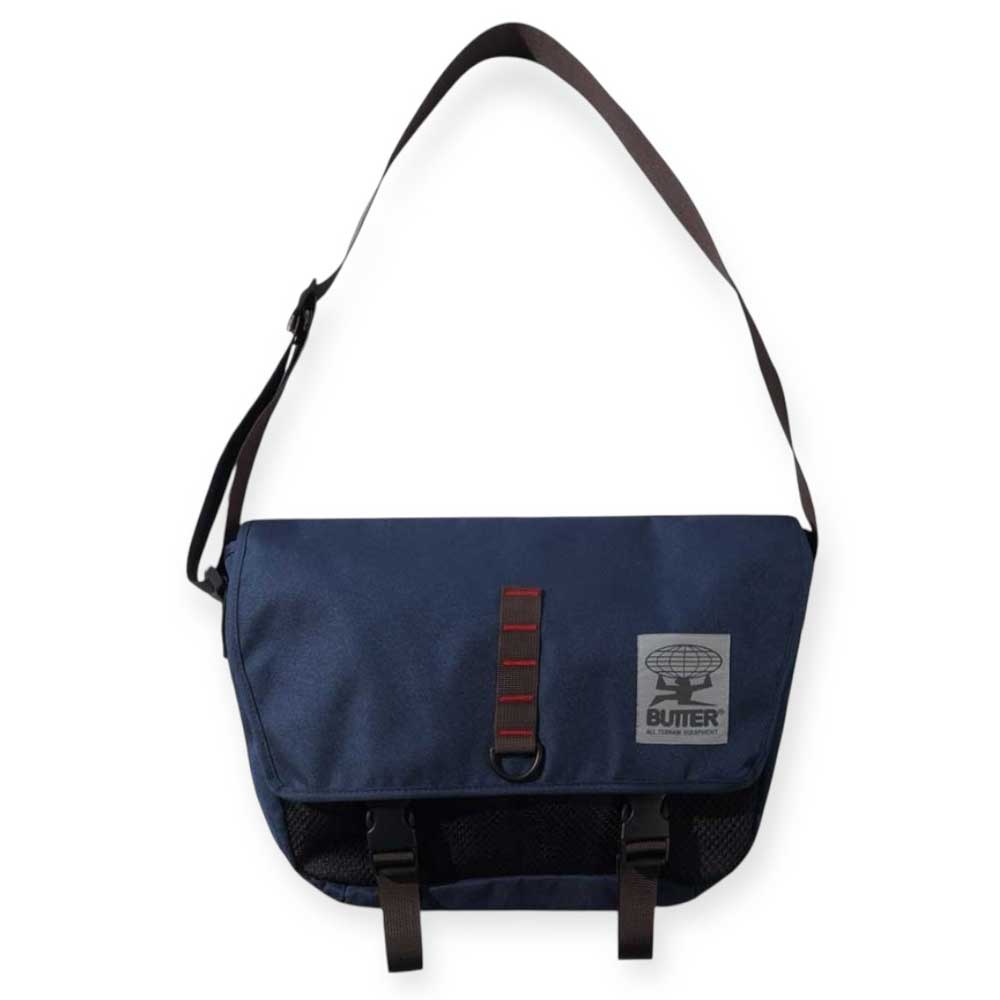 The Commute Side Bag Navy by Butter Goods is a 14L navy messenger bag in heavy cotton ripstop, featuring a black adjustable strap, stitched front accent, "BUTTER" mushroom patch, and two black buckle closures.