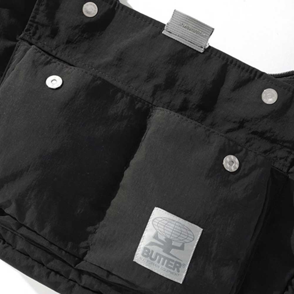 A close-up of the Crossbody Bag Black Butter Goods in heavy black cotton, featuring silver snap buttons, a gray fabric loop, and a gray label with a globe logo and "BUTLER" at the bottom.