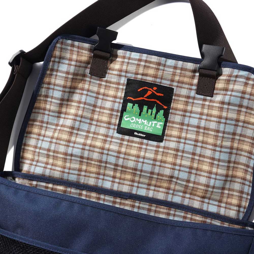 A close-up of the Butter Goods Crossbody Bag Sky Tan shows a blue flap, plaid interior with a COMMUTE CROSS BAG cityscape label, black adjustable straps, and sturdy clasps.