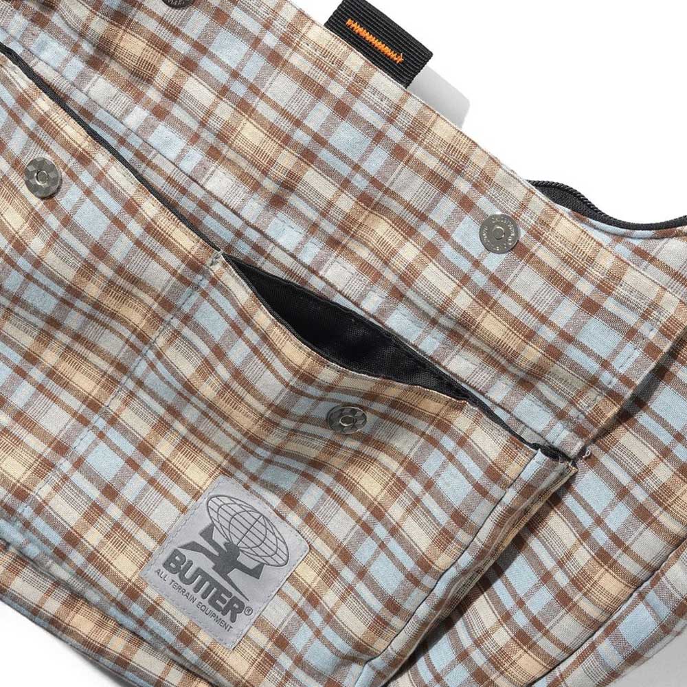 A close-up of the Crossbody Bag Sky Tan by Butter Goods in plaid beige, brown, and light blue cotton with buttons, a front pocket, and a stitched Butler logo patch featuring a globe graphic.