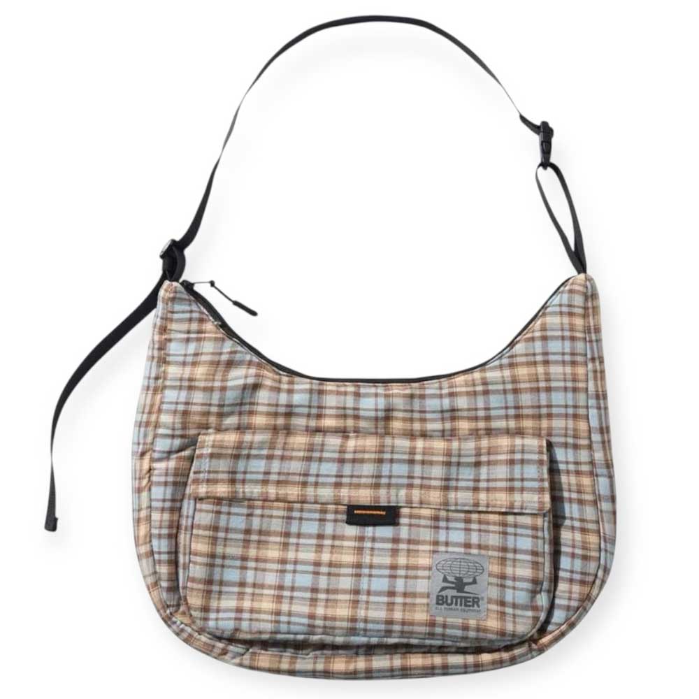 The Crossbody Bag Sky Tan by Butter Goods features a durable plaid ripstop design in brown, blue, and cream, an adjustable shoulder strap, front zip pocket, and a “BUTTER” patch with butter graphic.