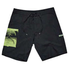 The Illusion 21 inch Trunk Black Former features black board shorts with a green abstract graphic on the left leg, a small white rectangular “Former” tag on the right leg, and a black drawstring at the waistband.