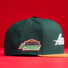 The El Tuga Houston Astros New Era 5950 Fitted hat features dark green with a brown brim, a colorful “Astrodome The Original” patch and stadium image on the side, and is shown resting on a red surface.