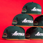 Five El Tuga Houston Astros New Era 5950 Fitted hats in dark green with orange brims are stacked against a bright red background. Each features the Astros logo and a stadium patch on the side.