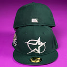 Two The Merc Green Silver 35th Houston Astros New Era Hat 5950 Fitted caps in dark green rest on a purple surface; the front displays a white star and oval, while the back shows a silver MLB logo above the closure.