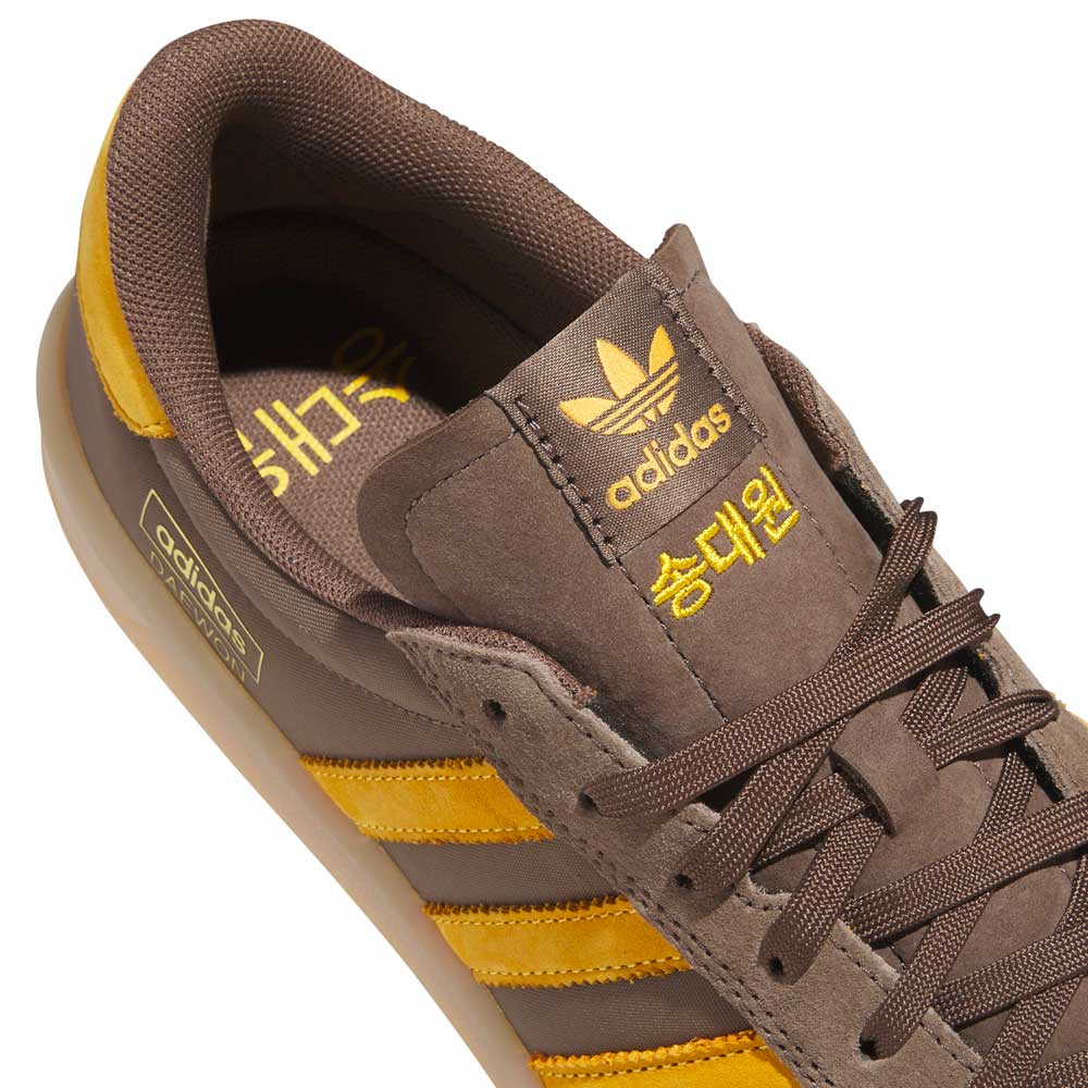 The Daewon Song Glenburn Brown Yellow KZ8777 Adidas Skateboarding Shoes feature a brown design with yellow stripes and logo, with Korean text displayed on the tongue. Captured from an angled overhead perspective, this street style sneaker highlights both its side and top details.