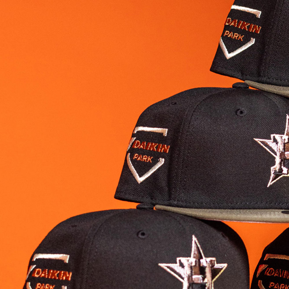 A close-up of three Daikin Park Olive Houston Astros New Era 5950 Fitted Hats stacked together, each featuring an orange and white H logo and Daikin Park embroidered on the side, set against an orange background.