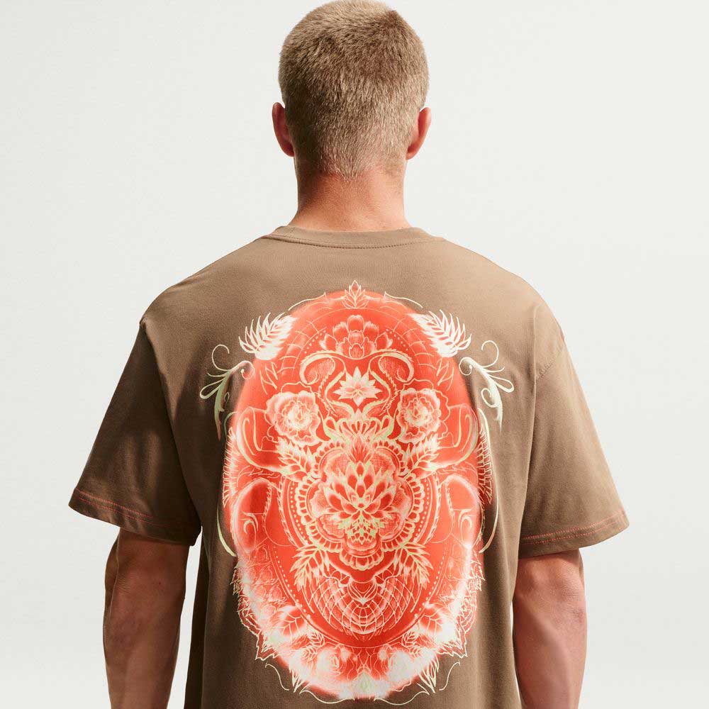 A person with short blond hair is seen from behind wearing the Nike SB Dark Driftwood Som Tum Dunk Tee (IH1478258), a roomy midweight cotton T-shirt with a large ornate orange and white floral graphic on the back, set against a plain light background.