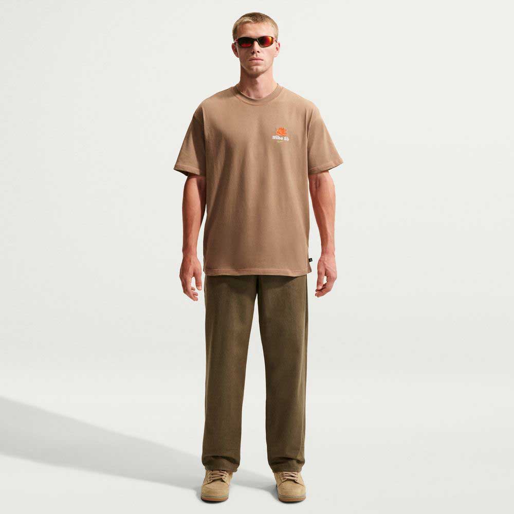 A man faces forward, wearing sunglasses and the Dark Driftwood Som Tum Dunk Tee IH1478258 by Nike SB Skateboarding, paired with olive green pants and beige shoes, set against a plain white background.