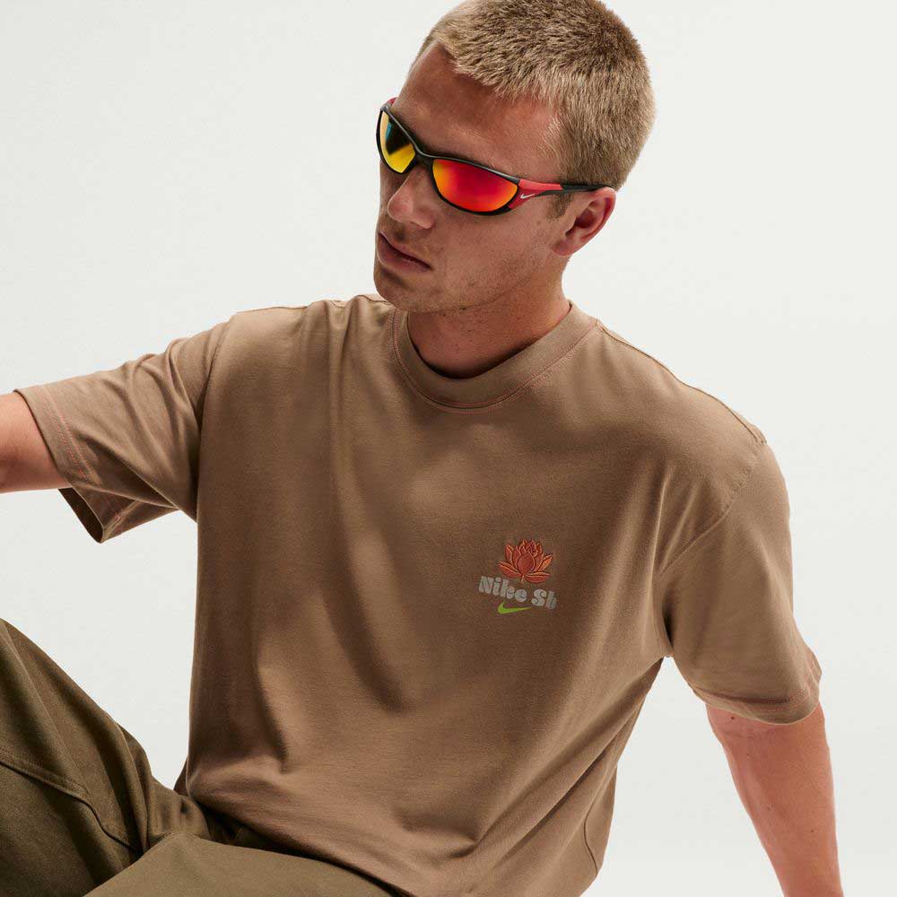 A man with short blonde hair wears red-tinted sunglasses and the Nike SB Dark Driftwood Som Tum Dunk Tee IH1478258, featuring a red flower and logo on the chest, paired with olive pants as he sits against a white background.