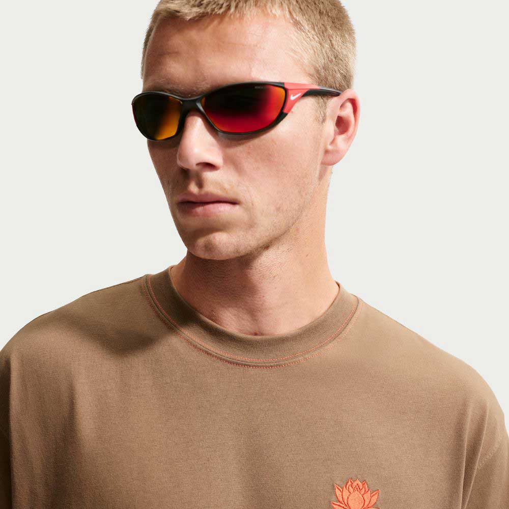 A man with short blond hair wears red-tinted sunglasses and the Nike SB Skateboarding Dark Driftwood Som Tum Dunk Tee, a brown midweight cotton shirt featuring an orange flower emblem on the chest, looking slightly to the side.