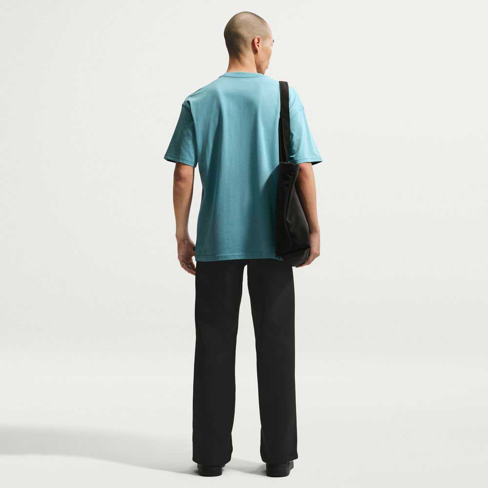 A person with a shaved head wears the Denim Turq Blue SF Muni Bus Pass Tee IU1265464 Nike SB Skateboarding T-shirt, black pants, and shoes, standing with a black shoulder bag against a plain white background.