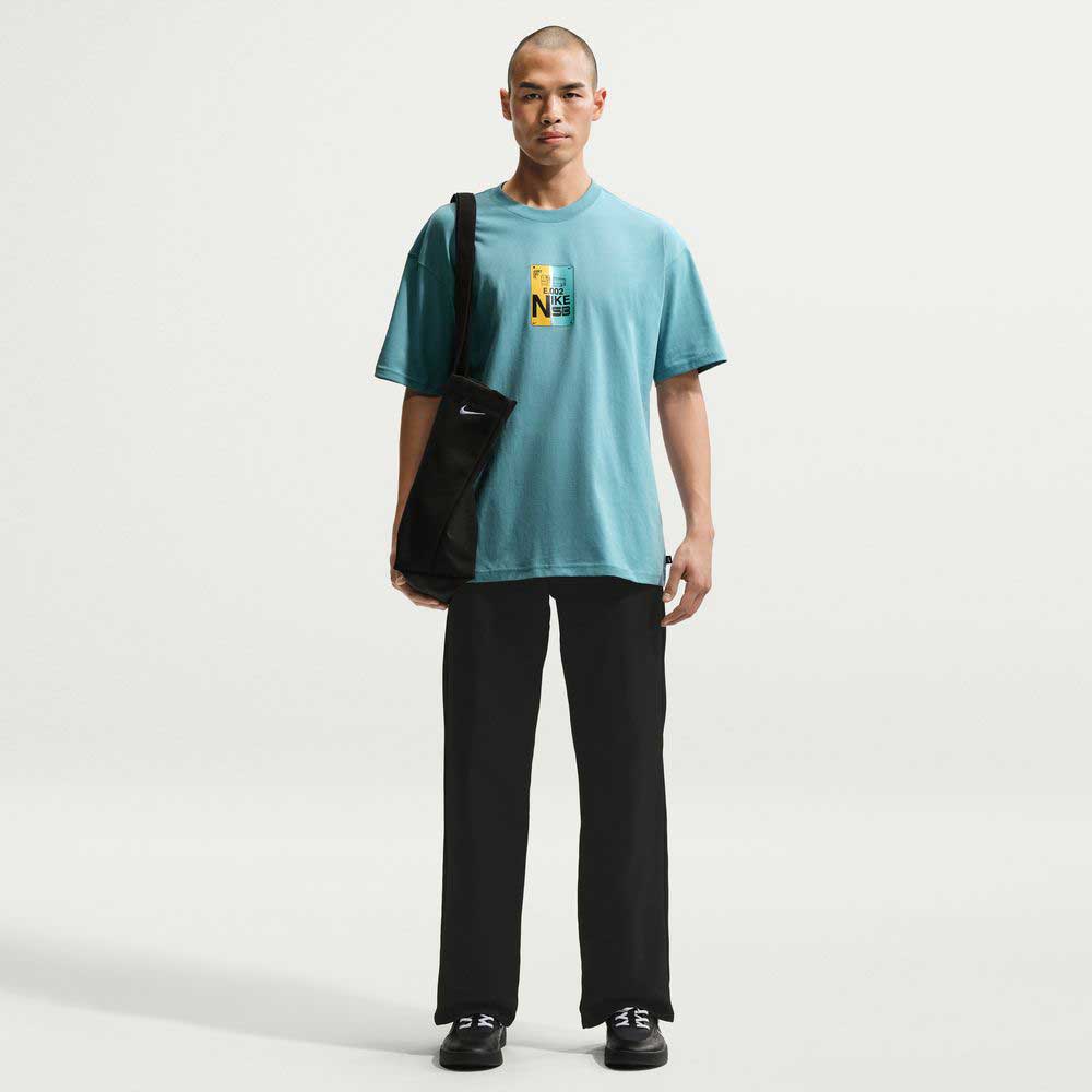 A person faces forward, wearing the Denim Turq Blue SF Muni Bus Pass Tee IU1265464 T-Shirt Nike SB Skateboarding Tee in light blue, black pants, black shoes, and a black shoulder bag against a plain white background.