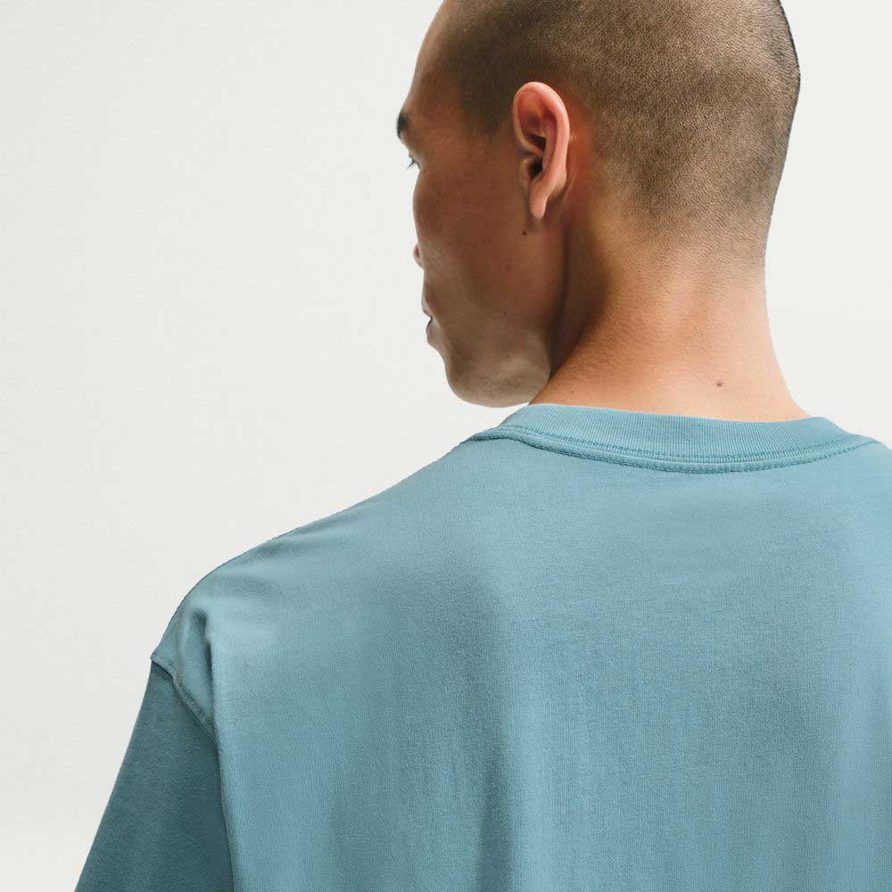 A person with short hair is shown from behind, wearing the Denim Turq Blue SF Muni Bus Pass Tee IU1265464 Nike SB Skateboarding T-Shirt in light blue against a plain, light background.