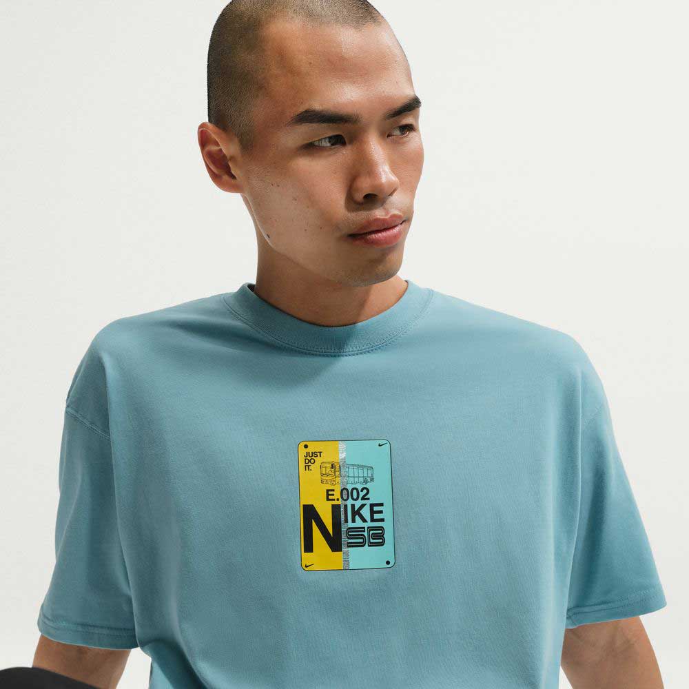 A person with a shaved head wears the Denim Turq Blue SF Muni Bus Pass Tee IU1265464, a Nike SB Skateboarding midweight cotton T-shirt in light blue, featuring a multicolored chest patch, shown against a plain white background.