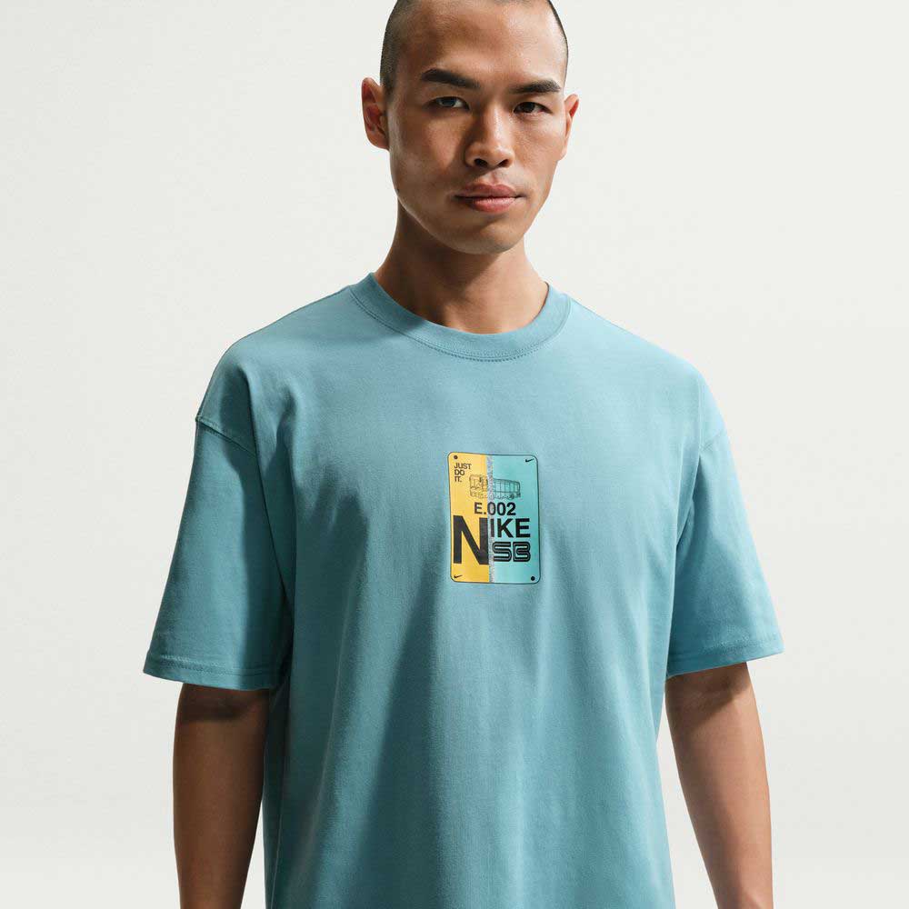 A person wears the Nike SB Denim Turq Blue SF Muni Bus Pass Tee (IU1265464), a laid-back midweight cotton tee with a colorful graphic, against a plain white background.