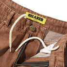Close-up of Distressed Brown Relaxed Denim Jeans by Butter Goods, made from 100% cotton with an internal drawstring waistband, metal button, and yellow RELAXED LOOSE FIT tag; the zipper is partially open.